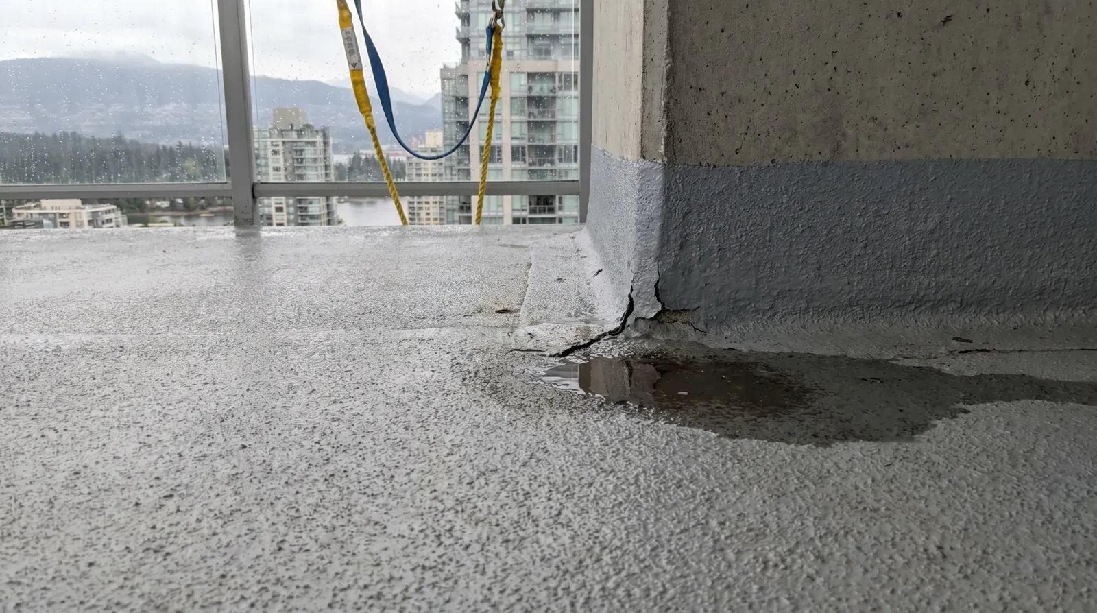 Failed liquid polyurethane traffic-coating membrane on a Vancouver strata high-rise balcony deck with water ponding at a cracked wall-to-floor upturn, glass railing and rainy coastal skyline behind.